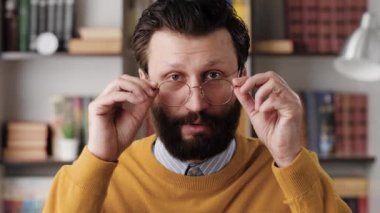 Man is surprised, shocked. Confused bearded man in glasses in office or apartment room looking at camera and slowly takes off his glasses in surprise looking at camera. Close up and slow motion