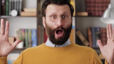 Man is surprised, shocked. Smiling bearded man in glasses in office or apartment room looking at camera and waves his hands emotionally expressing his incredible surprise. Close up and slow motion