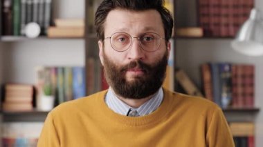 Shrug, i dont know. Perplexed bearded man in glasses in office or apartment room looking at camera and emotionally squeezes his shoulders and spreads his hands to show his ignorance. Close up