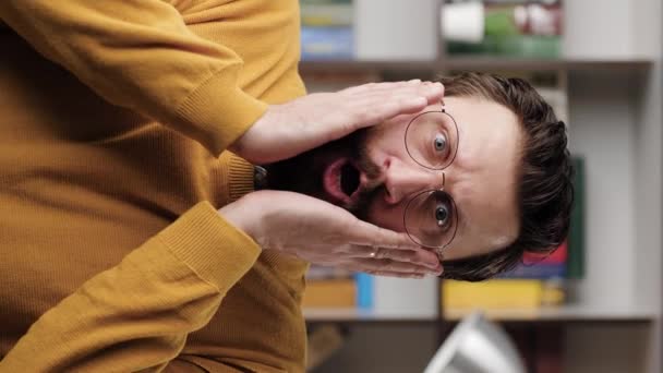 Man is surprised, shocked. Vertical video of smiling bearded man in glasses in office or apartment room looking at camera and waves his hands emotionally expressing his incredible surprise. Close up — Wideo stockowe