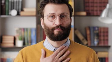 Respect bow. Bearded man with glasses in office or apartment room looking at camera and puts his hand on his chest and bows, expressing respect or greeting. Close-up and slow motion