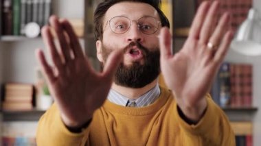 Disagreement, stop, NO says. Frightened worried bearded man in glasses in office or apartment room looks at camera and starts waving his arms nervously, shouting no-no-no. Close-up and slow motion