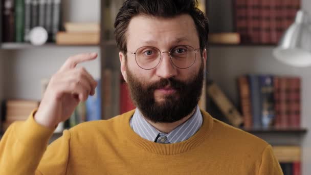 Estúpida. Hombre barbudo irritado en gafas en la oficina o habitación del apartamento mira a la cámara y gira su dedo en su sien. Primer plano y cámara lenta — Vídeos de Stock