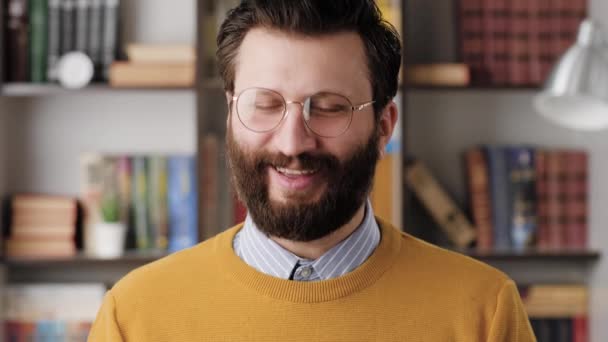 Человек улыбается, смеется. позитивный веселый бородатый мужчина в очках в офисе или в квартире смотрит в камеру, улыбается и робко смеется. крупный план и замедленная съемка — стоковое видео