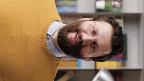 Man smiling, laughs. Vertical video of positive joyful bearded man in glasses in office or apartment room looking at camera and smiles and laughs shyly. Close-up and slow motion — Wideo stockowe