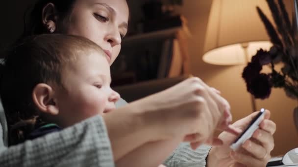 Mama i dziecko bawią się przez telefon. Wieczorem, siedzi przy stole w sypialni kobieta trzyma słodkie dziecko 2-3 lat na kolanach i naciska go palcem na ekranie smartfona. Zbliżenie i spowolnienie ruchu — Wideo stockowe