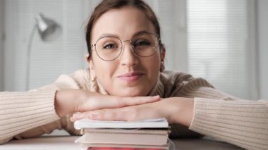 Yabancı eğitim, uzaktan eğitim, çevrimiçi eğitim, uzaktan sunum kavramları. Gözlüklü gülümseyen öğrenci kadın kitap yığınına yaslanmış bilgisayar kamerasına bakıyor ve göz kırpıyor.