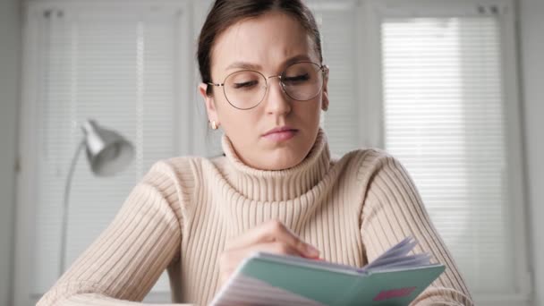 Online learning, online courses concept. Student woman with glasses sits in front of computer webcam talking and takes written notes, she looking at camera and maintains internet conversation — Stock Video