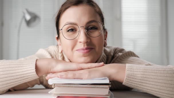 Buitenlands onderwijs, afstandsonderwijs, online onderwijs, concepten voor presentatie op afstand. Lachende student vrouw met bril leunend op stapel boeken kijken naar computer camera en knipogen — Stockvideo