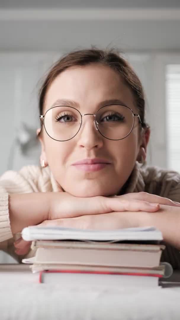 Educação estrangeira, educação a distância, educação on-line, conceitos de apresentação remota. Vídeo vertical de mulher estudante sorrindo apoiando-se na pilha de livros olhando para a câmera do computador e piscando — Vídeo de Stock