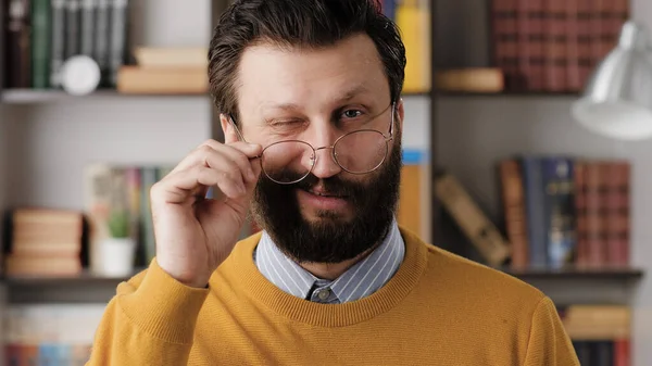 Man winks. Positive smiling bearded man with glasses in office or apartment room looking at camera and slowly lowers his glasses and winks. Close up view