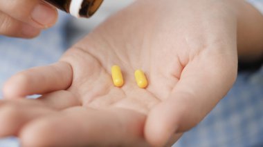 Taking pills. Two small bright yellow cylindrical pill capsules fall into palm of hand from pill bottle. Close-up, front view, center composition