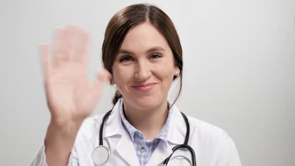 Docteur vous salue. Femme médecin souriante positive sur fond gris regardant la caméra et agitant la main droite montrant salutation. Mouvement lent — Video