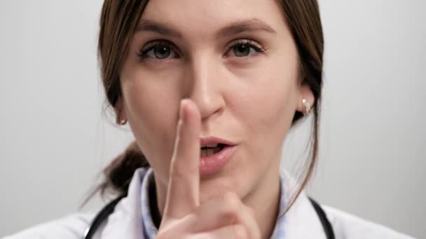 Dedo secreto del doctor. Mujer doctora sospechosa sobre fondo gris mirando a la cámara y trae su dedo índice a los labios de su boca y dice shhh. Movimiento lento — Vídeo de stock