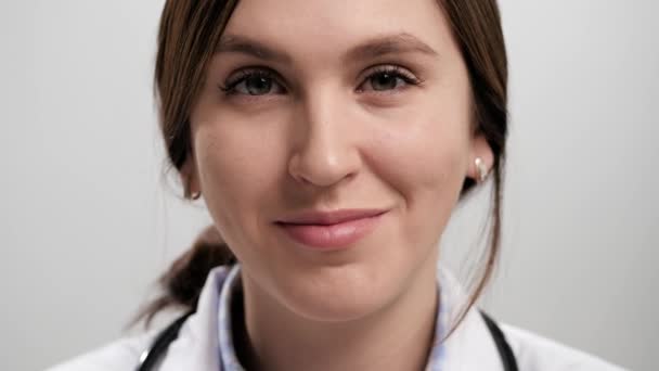 O doutor pisca. Close-up de sorrindo positivo médico mulher no fundo cinza olhando para a câmera e brincando piscando com o olho direito. Movimento lento — Vídeo de Stock