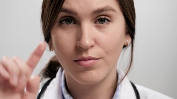 Dokter menunjukkan kebodohan. Close-up of iritasi perempuan dokter pada latar belakang abu-abu melihat kamera dan memutar jarinya di pelipisnya. Gerakan lambat — Stok Video