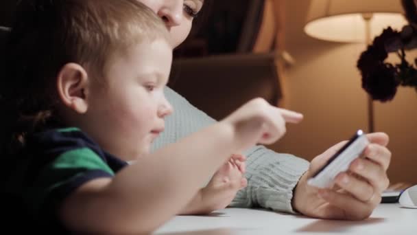 El bebé juega con el teléfono. La mujer sostiene el teléfono en su mano y su hijo pequeño se sienta cerca y toca en la pantalla del teléfono inteligente, la madre le dice algo al bebé y está feliz por su éxito. Primer plano cámara lenta — Vídeos de Stock