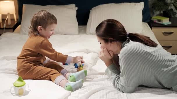 Mamá y el bebé están jugando. Mujer y niño de 2-3 años en pijama se acuestan en la cama cara a cara por la noche y juegan con juguete educativo, se ríen y se regocijan. Movimiento lento — Vídeo de stock