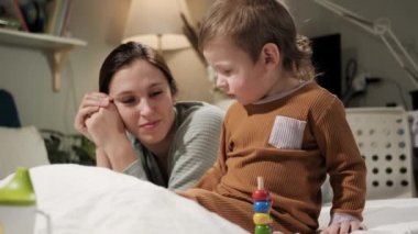 Bebek oynuyor, anne çocuğuna hayran. 2-3 yaşlarında mutlu bir kadın ve erkek çocuğu akşamları pijamalarıyla yatağa uzanır ve çocuk eğitici oyuncaklarla oynar, anne oğlunu ilgiyle izler. Yavaş çekim