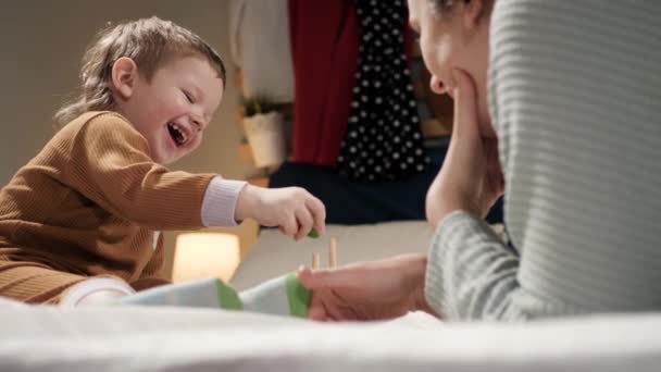 Máma a dítě si hrají. Pohled na malého veselého smějícího se chlapce 2-3 roky, který leží za zády své matky, večer leží v pyžamu na posteli a hrají si se vzdělávací hračkou. Zpomalený pohyb — Stock video
