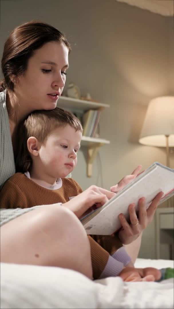 Mãe e bebê livro de leitura. Vista vertical lateral da mulher e menino de 2-3 anos de idade sentar na cama de pijama no quarto à noite ou à noite e segurar livro em mãos, olhando e lendo. Movimento lento — Vídeo de Stock