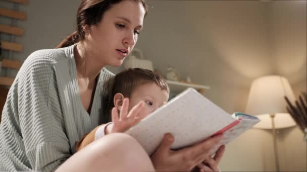 Baby and book, mother and child reading. Woman mom and 2-3 year old boy sit on bed in pajamas in bedroom in evening or night and hold book in hands, looking and reading, turn page. Slow motion — Stock Video