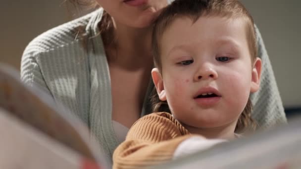 Mother and baby reading book. Front view of face of 2-3 year old baby who sits with his mother on bed, female hands hold book and boy looks at it or reads it carefully. Slow motion and close-up — Stock Video