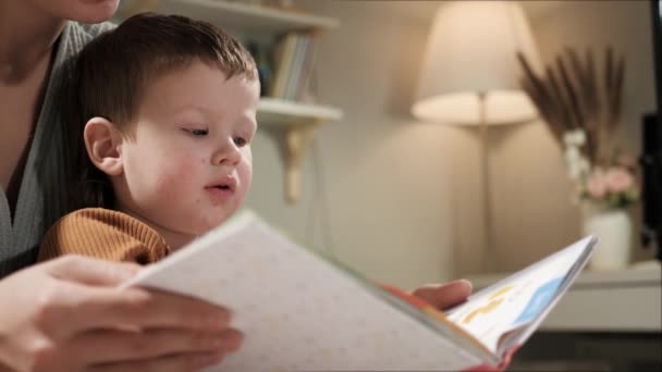 Bayi dan buku. Pandangan samping dari wajah bayi 2-3 tahun yang duduk bersama ibunya di tempat tidur di kamar tidur, tangan wanita memegang buku dan anak laki-laki melihatnya atau membacanya dengan hati-hati. Gerakan lambat — Stok Video