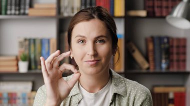 Kadın iyi görünüyor. Ofiste ya da dairede gülümseyen olumlu bir kadın kameraya bakar ve parmaklarıyla iyi bir hareket gösterir. Orta ekran görüntü