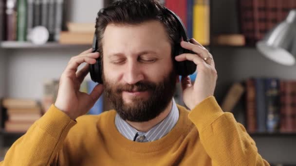 El hombre con auriculares escucha música. Hombre barbudo positivo con auriculares inalámbricos negros sostiene su cabeza con las manos y disfruta de la música. Movimiento lento — Vídeo de stock
