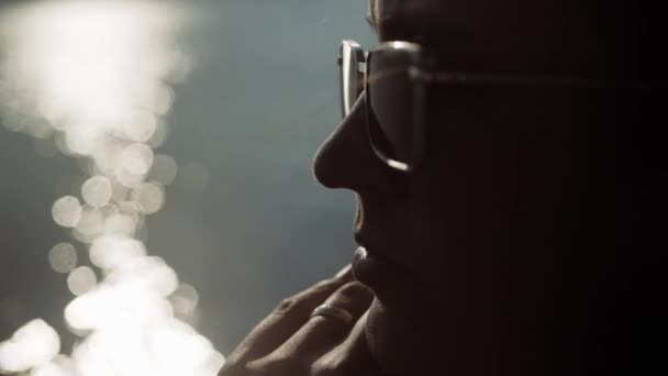 Frau mit Sonnenbrille denkt nach. Großaufnahme der Schönheit Frauengesicht mit Sonnenbrille, die nachdenklich nach irgendwo bei Sonnenuntergang schaut, im Hintergrund Wasser mit gleißendem Sonnenlicht. Zeitlupe — Stockvideo