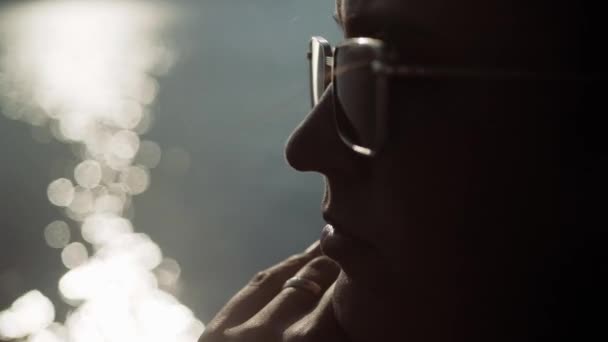 Mujer con gafas de sol pensando. Primer plano de la cara de la mujer de belleza en gafas de sol que está mirando pensativamente a algún lugar al atardecer, en el agua de fondo con el resplandor del sol. Movimiento lento — Vídeos de Stock