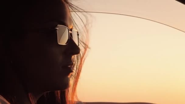 La mujer conduce el coche. Mujer hermosa feliz en gafas de sol conduce coche con ventana abierta al atardecer, el viento vuela maravillosamente su cabello. Primer plano — Vídeos de Stock