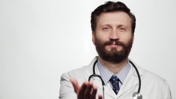 Doktor avucuyla işaret ediyor. Beyaz arka planda gülümseyen doktor portresi kameraya bakıyor ve işaret ediyor, avucuyla işaret ediyor. — Stok video