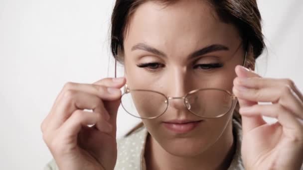 Kvinnan tar på sig glasögon. Positiv kvinna på vit bakgrund sätter på glasögon och leenden medan du tittar på kameran. Vision, student-, lärar- och utbildningskoncept — Stockvideo