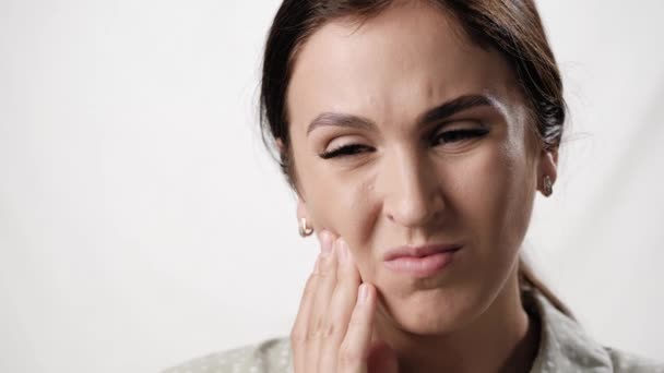 Οδοντόκρεμα. Πρόσωπο μιας γυναίκας που υποφέρει με πονόδοντο σε λευκό φόντο, αγγίζει το μάγουλό της με το χέρι της προσπαθώντας να ανακουφίσει τον πόνο των δοντιών. Κοντινό πλάνο και αργή κίνηση — Αρχείο Βίντεο