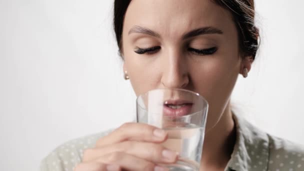 A nő vizet iszik. A fehér hátterű nő üvegből issza a vizet, és mosollyal néz a kamerába. Közelkép és lassított felvétel