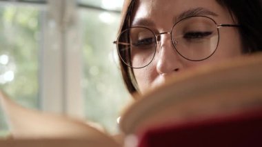 Kitap okuyan ve kahve içen bir kadın. Gözlüklü kadın kitap okuyor ve fincandan kahve ya da çay içiyor. Yakın çekim ve yavaş çekim