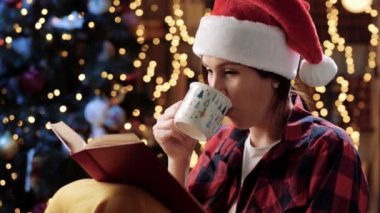 Noel konsepti. Kırmızı şapkalı güzel bir kadın oturmuş kitap okuyor ve fincandan bir şeyler içiyor. Arka planda çelenklerin ve Noel ağacının bulanık ışıkları.