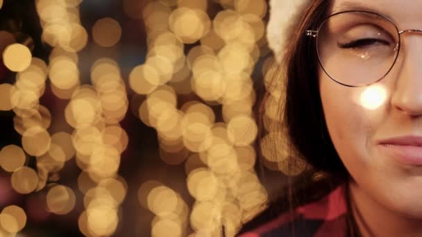 Boldog karácsonyt és boldog új évet! Közelkép egy félarcú nőről, aki a kamerába néz. A szemüveg karácsonyi fényeket és csillagszórókat tükröz homályos fények mögött. — Stock videók