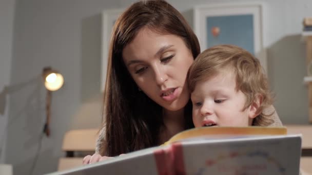 Mamá y el bebé están leyendo. Vista frontal de las caras de la madre y el niño que están sentados con el libro, la lectura. Primer plano y cámara lenta — Vídeo de stock