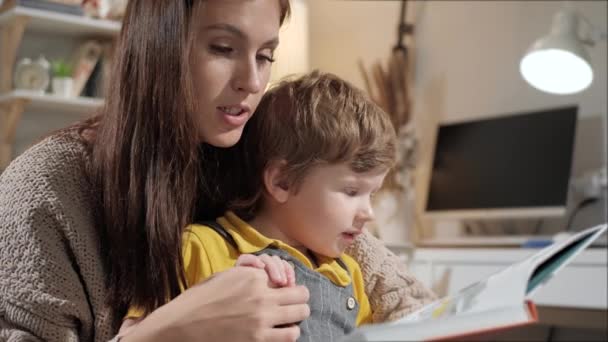 Annem bebeğe kitap okuyor. Anne ve çocuk akşam yatak odasında yatakta oturuyor, anne oğlanın elini tutuyor ve ona kitap okuyor. Yakın çekim ve yavaş çekim — Stok video