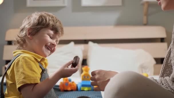 Baby eet snoep. Gelukkig lachend lachend kind zit op bed in de slaapkamer en eet chocolade snoep en trakteert haar op mama. Close-up en slow motion — Stockvideo