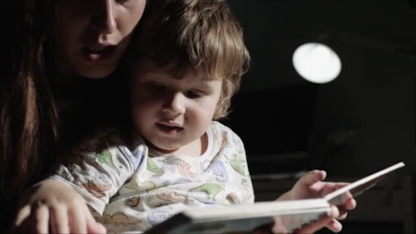 Мама и ребенок читают книгу. Мама читает книгу ребенку перед сном, ребенок слушает и смеется. Крупный план и замедленная съемка — стоковое видео