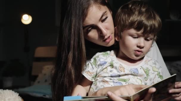 Мать и ребенок читают книгу. Мама и ребенок 2-3 лет в пижаме сидят ночью на кровати в спальне и читают книги. Крупный план и замедленная съемка — стоковое видео