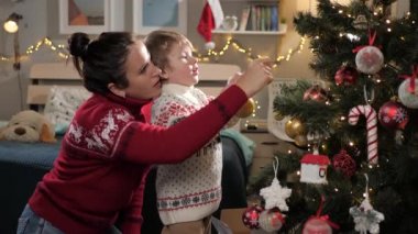Anne ve çocuk Noel ağacı süslüyorlar. 2-3 yaşlarındaki anne ve çocuğu Noel ağacını süslüyor, dallara oyuncak takıyor. Arka planda Noel ışıkları parlıyor. Yavaş çekim