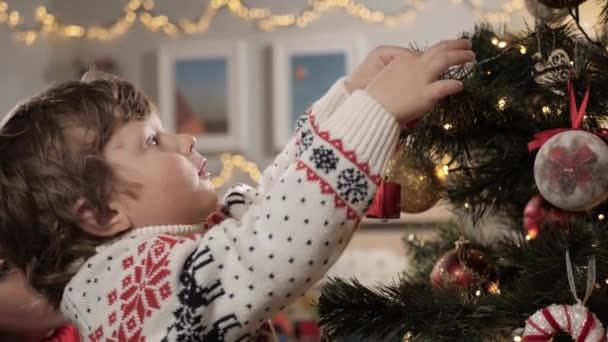 Grabben sätter leksak på julgranen. Pensiv barn dekorerar julgran och noggrant hänger leksak på gren, i bakgrunden mamma tittar på baby. Närbild och slow motion — Stockvideo