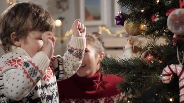 Kind schmückt Weihnachtsbaum. Glückliches Kind legt Spielzeug auf den Weihnachtsbaum und freut sich sehr über diesen Moment, Mutter im Hintergrund schaut Baby an und lächelt. Nahaufnahme und Zeitlupe — Stockvideo