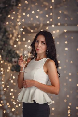 Noel ve yeni yıl büyüsü. Peri güzelliğindeki Noel arifesinde şenlikli bir gece elbisesi içinde bir bardak şampanyası olan büyüleyici, mutlu genç bir kadın. Işıklar yansın.