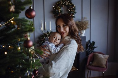 Beyaz kazaklı anne küçük çocuğu öpüyor Noel ağacındaki süveterli çocuğu ve evdeki çelenkleri.